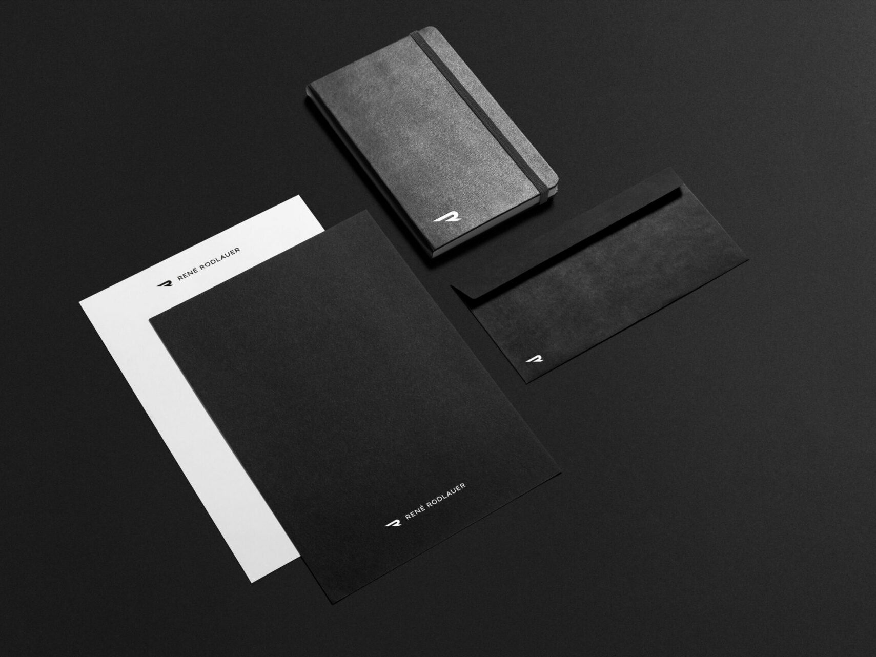 Briefpapier, Notizbuch und Briefumschlag mit Icon sowie Logo von René Rodlauer auf schwarzem Hintergrund.