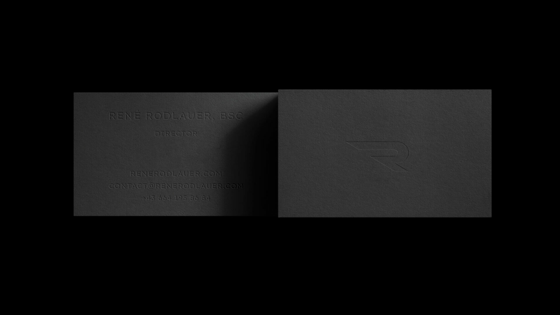 Graue Vorder- und Hinterseite der René Rodlauer Visitenkarte auf schwarzem Hintergrund. Auf der Vorderseite geprägt ist das R-Icon. Auf der Rückseite geprägt sind die Kontaktdaten.