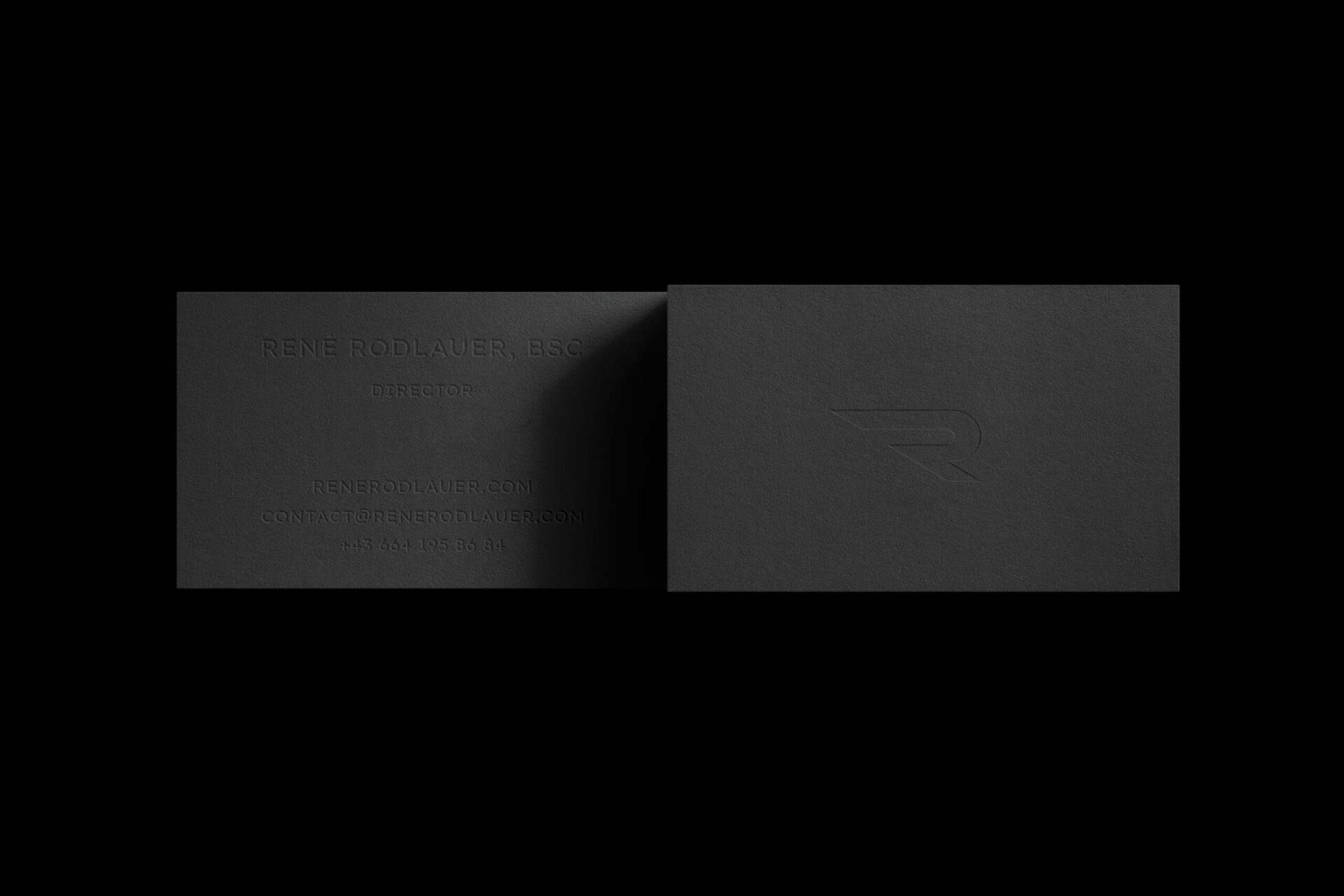 Graue Vorder- und Hinterseite der René Rodlauer Visitenkarte auf schwarzem Hintergrund. Auf der Vorderseite geprägt ist das R-Icon. Auf der Rückseite geprägt sind die Kontaktdaten.